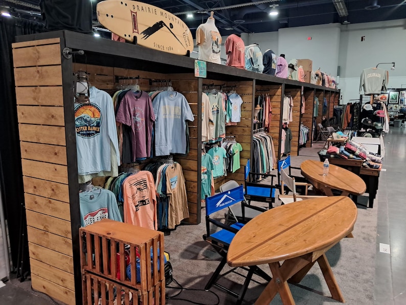 May include: A retail display booth featuring wooden shelving units filled with various graphic t-shirts, a surfboard-shaped sign labeled Prairie Mountain, and two surfboard-shaped wooden tables with blue director chairs in a large indoor venue.