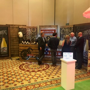 May include: A promotional trade show booth for Harken wines featuring wooden panels, decorative bicycle wheel accents, and large black signage with the text TAKE A RIDE. A white pedestal displays a light box with the text #LIVE FOR THE DAY!