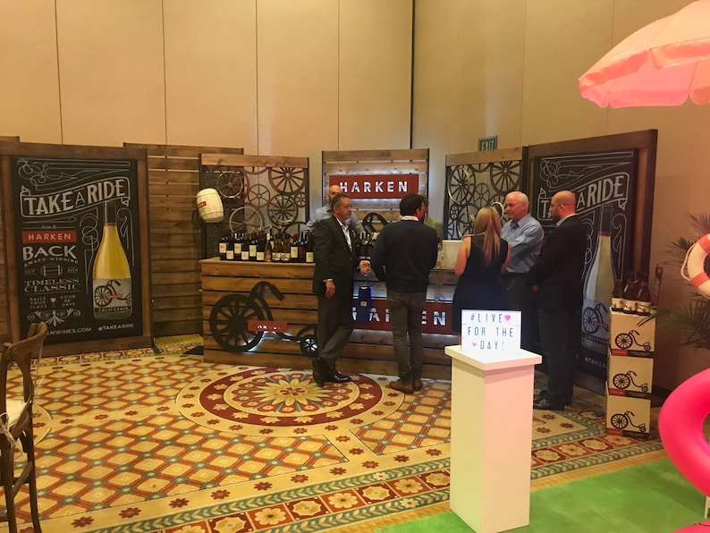 May include: A promotional trade show booth for Harken wines featuring wooden panels, decorative bicycle wheel accents, and large black signage with the text TAKE A RIDE. A white pedestal displays a light box with the text #LIVE FOR THE DAY!
