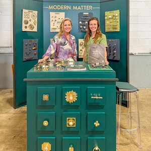May include: A dark green display booth labeled MODERN MATTER featuring a collection of gold-toned cabinet hardware, including various knobs, pulls, and handles mounted on boards and a central cabinet unit with nine drawers displaying decorative hardware.