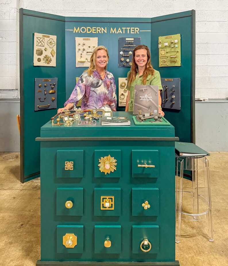 May include: A dark green display booth labeled MODERN MATTER featuring a collection of gold-toned cabinet hardware, including various knobs, pulls, and handles mounted on boards and a central cabinet unit with nine drawers displaying decorative hardware.