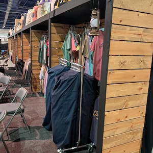 May include: A retail display structure made of horizontal light wood planks and dark metal framing. The unit features hanging racks filled with various t-shirts, a rolling garment rack covered in dark blue fabric, and a vintage-style metal lantern.