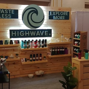 May include: A trade show display featuring a light wood paneled wall with a circular wave logo and the text HIGHWAVE, WASTE LESS, and EXPLORE MORE. Multiple shelves display various colorful reusable water bottles and pet bowls in a retail setting.