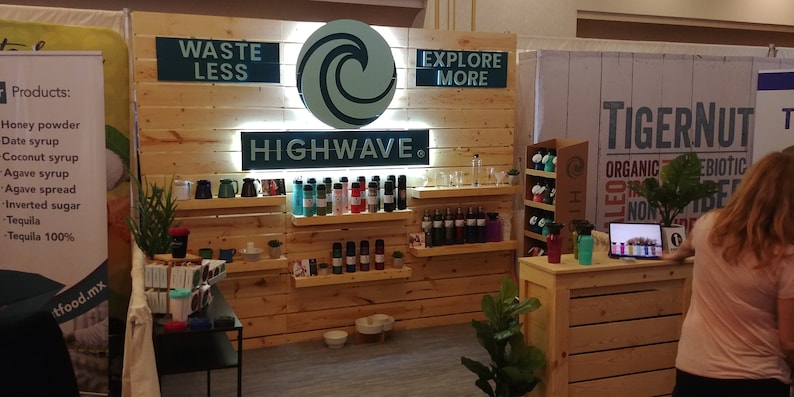 May include: A trade show display featuring a light wood paneled wall with a circular wave logo and the text HIGHWAVE, WASTE LESS, and EXPLORE MORE. Multiple shelves display various colorful reusable water bottles and pet bowls in a retail setting.