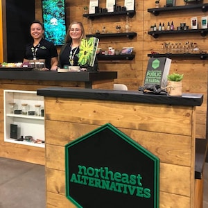May include: A wooden retail counter featuring a black hexagonal sign with the green text Northeast Alternatives. A sign on the counter reads Public Consumption Is Illegal. Shelves behind the counter display various products and small potted plants.