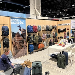 May include: A trade show booth display featuring various Eddie Bauer backpacks and luggage in colors including black, blue, red, green, and tan. The display includes wall-mounted bags, a large branded poster, and a white table with rolling suitcases.
