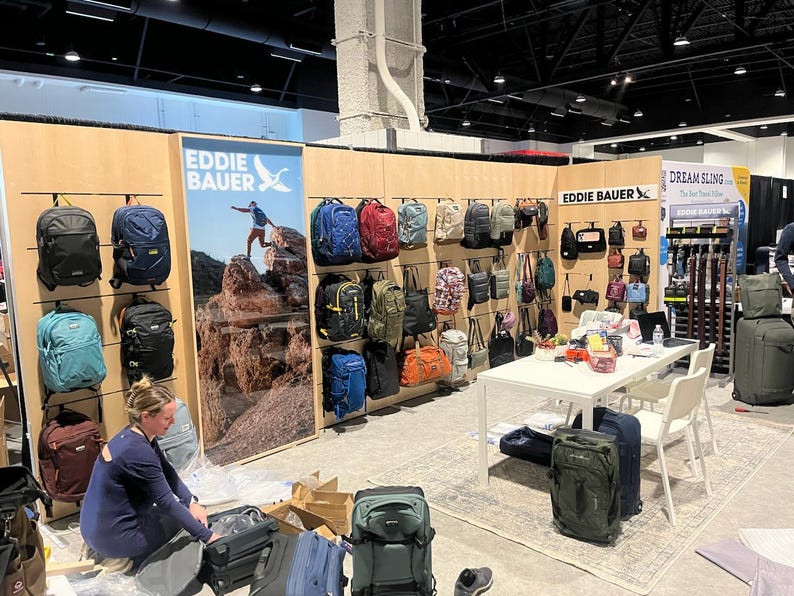 May include: A trade show booth display featuring various Eddie Bauer backpacks and luggage in colors including black, blue, red, green, and tan. The display includes wall-mounted bags, a large branded poster, and a white table with rolling suitcases.