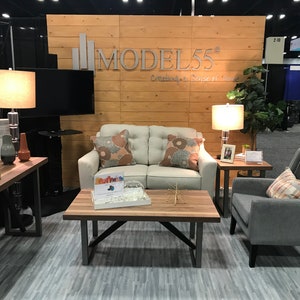 May include: A living room display featuring a cream-colored loveseat with two patterned pillows, a gray armchair with a plaid pillow, a wooden coffee table, a side table with a lamp, and a console table with a lamp, all set against a wood-paneled wall.