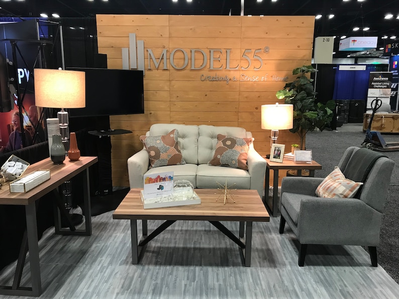 May include: A living room display featuring a cream-colored loveseat with two patterned pillows, a gray armchair with a plaid pillow, a wooden coffee table, a side table with a lamp, and a console table with a lamp, all set against a wood-paneled wall.