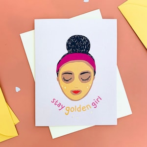 Può includere: Un biglietto d'auguri con un'illustrazione rosa e oro di una persona con una maschera per il viso. Il testo sulla carta dice "stay golden girl".