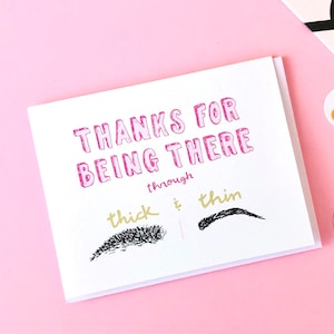 May include: A white greeting card with the text "THANKS FOR BEING THERE" in pink, and "through thick & thin" in gold. The card features illustrations of two eyebrows, one thick and one thin, on a pink background.
