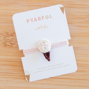 May include: A white and pink ice cream cone charm with a pink string attached. The charm is handmade by women artisans in Uttarakhand, India through Himalayan Blooms. The card reads "Pyarful Rakhi".