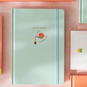 May include: A light green journal with a blue ribbon closure. The cover features a gold illustration of a teapot pouring tea into a cup with the text "WRITE STUFF & drink chai".