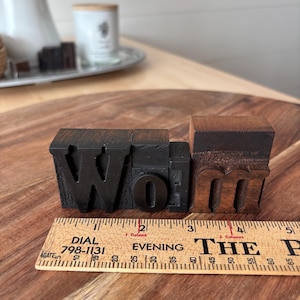 May include: Wooden letterpress printing blocks spelling "Wolm" in a dark color. A wooden ruler with the text "DIAL 798-1131 EVENING THE P" is placed below the blocks. The blocks are on a wooden surface.
