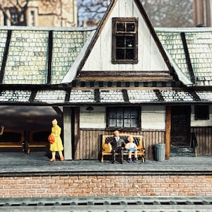 May include: Miniature model of a train station with a brick base, a sloped roof, and a waiting area with benches. Small figures of people are placed around the station. The roof is detailed with a textured pattern.