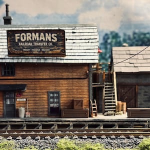 May include: A miniature model of a wooden building labeled Formans Railroad Transfer Co. with a shingled roof, a loading dock, stairs, and crates. The structure features a brown exterior, a small office door, and a utility pole with a wire.