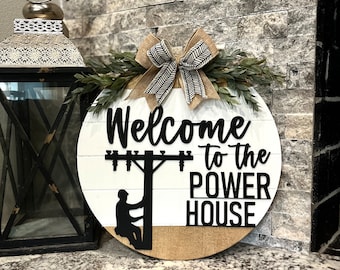 Puede incluir: Cartel de madera redondo con el texto "Welcome to the Power House". El cartel presenta una silueta de un liniero en un poste de energía. Está decorado con un lazo de arpillera y vegetación.