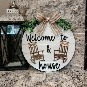 May include: Welcome to our house sign with a brown burlap bow and green leaves. The sign features two rocking chairs with the words "giga" and "papa" on them.