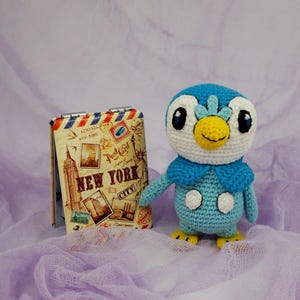 May include: A blue and white crocheted penguin plush toy with a yellow beak and black eyes. The toy is sitting on a purple and white fabric background. The toy is next to a small vintage-style notebook with the words "New York City" printed on the cover.
