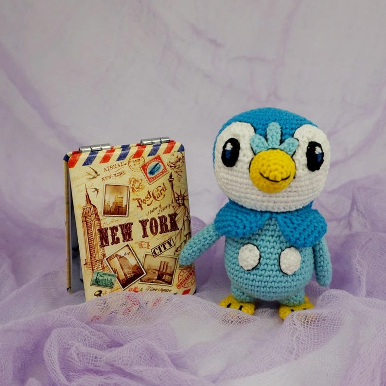 May include: A blue and white crocheted penguin plush toy with a yellow beak and black eyes. The toy is sitting on a purple and white fabric background. The toy is next to a small vintage-style notebook with the words "New York City" printed on the cover.