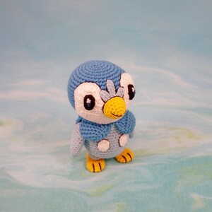May include: A blue and white crocheted penguin plush toy with yellow beak and feet. The penguin has black eyes and a white face.
