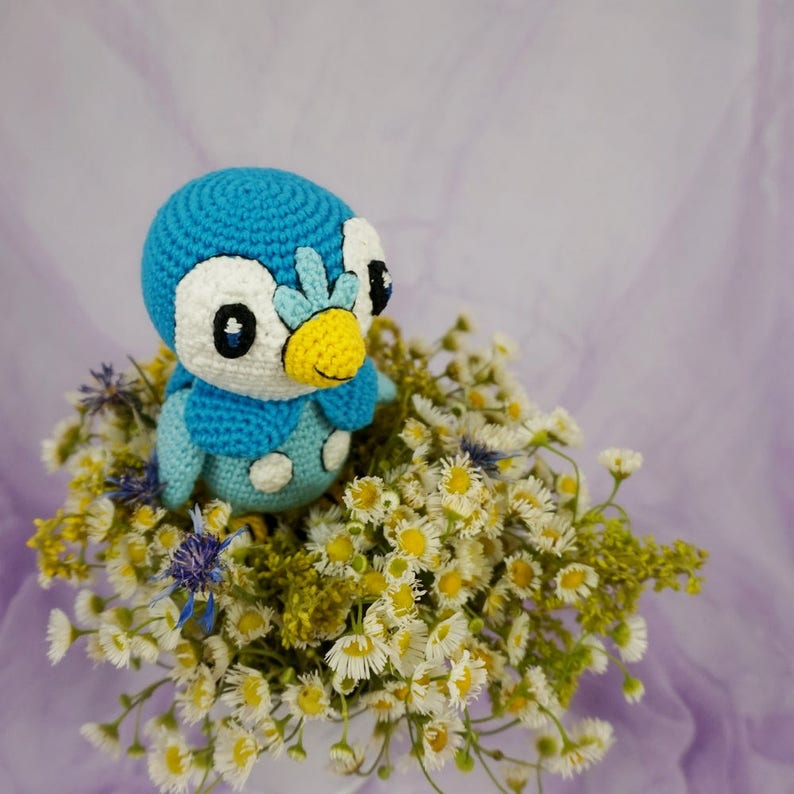 May include: A blue and white crocheted penguin toy sitting in a bouquet of white daisies and yellow wildflowers.