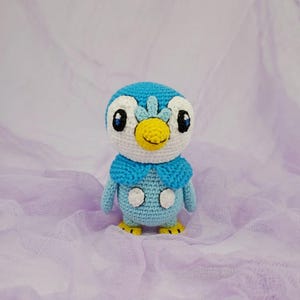 May include: A blue and white crocheted penguin plush toy with a yellow beak and black eyes. The penguin is wearing a blue hooded scarf.