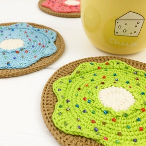 May include: Three crocheted donut coasters in green, blue, and pink with white centers and colorful sprinkles. The coasters are on a white surface.