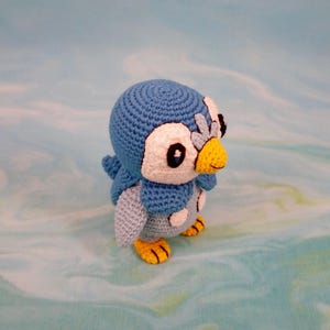 May include: A blue and white crocheted penguin plush toy with yellow beak and feet. The penguin is standing on a blue and white background.