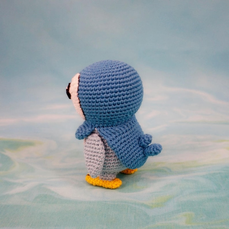 May include: A blue crocheted penguin toy with yellow feet. The penguin is facing away from the camera and is standing on a light blue surface.