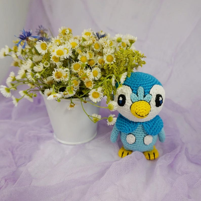 May include: A blue and white crocheted penguin plush toy with a yellow beak and white eyes. The toy is sitting on a light purple fabric background with a bouquet of white daisies in a white bucket.
