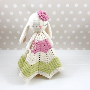Puede incluir: Un juguete de crochet en forma de conejo con una flor rosa en la cabeza, sosteniendo una manta de crochet multicolor. La manta es blanca, rosa y verde.