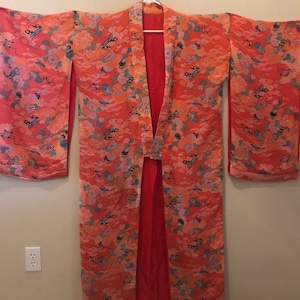 Pode incluir: Um quimono comprido, vermelho e laranja com um padrão floral. O quimono tem um colar largo e aberto e mangas compridas. O tecido é sedoso e tem um brilho sutil.
