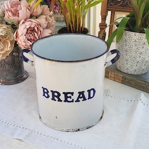 May include: White enamel bread bin with blue trim and the word "BREAD" in blue lettering. The bin is sitting on a white tablecloth with a floral pattern.