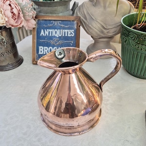 May include: A polished copper jug with a handle and spout, reflecting the light, is placed on a white tablecloth. The background includes flowers, a bust, and a sign that reads "ANTIQUITES BROC".