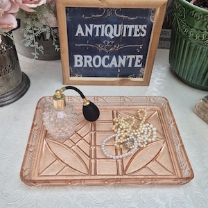 May include: A pink glass tray with a geometric pattern, displaying a perfume bottle with a black atomizer and pearl necklaces. A framed sign reads "ANTIQUITES BROCANTE".