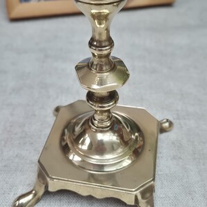 Peut inclure: Un bougeoir en laiton poli dot&eacute; d'une base carr&eacute;e avec quatre pieds d&eacute;coratifs et une tige centrale compos&eacute;e de sections &eacute;tag&eacute;es, arrondies et hexagonales. La surface m&eacute;tallique est r&eacute;fl&eacute;chissante et lisse, avec une finition dor&eacute;e.