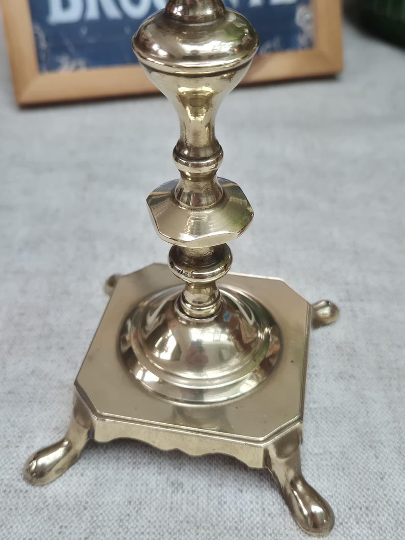 Peut inclure: Un bougeoir en laiton poli dot&eacute; d'une base carr&eacute;e avec quatre pieds d&eacute;coratifs et une tige centrale compos&eacute;e de sections &eacute;tag&eacute;es, arrondies et hexagonales. La surface m&eacute;tallique est r&eacute;fl&eacute;chissante et lisse, avec une finition dor&eacute;e.