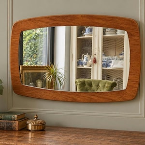 May include: A rectangular wall mirror with rounded corners and a medium-toned wood grain frame. The mirror features a smooth, polished surface that reflects a room interior, including a white cabinet, a green chair, and a window with greenery.
