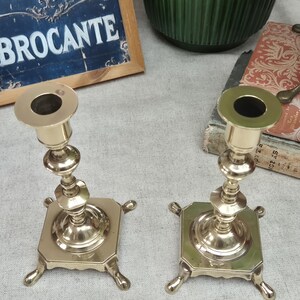 Peut inclure: Une paire de bougeoirs en laiton poli dot&eacute;s d'une tige centrale avec des d&eacute;tails tourn&eacute;s d&eacute;coratifs, d'une coupelle sup&eacute;rieure circulaire et d'une base carr&eacute;e soutenue par quatre petits pieds incurv&eacute;s &agrave; chaque coin de la base m&eacute;tallique.