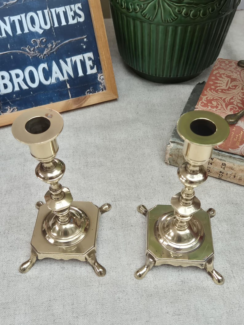 Peut inclure: Une paire de bougeoirs en laiton poli dot&eacute;s d'une tige centrale avec des d&eacute;tails tourn&eacute;s d&eacute;coratifs, d'une coupelle sup&eacute;rieure circulaire et d'une base carr&eacute;e soutenue par quatre petits pieds incurv&eacute;s &agrave; chaque coin de la base m&eacute;tallique.