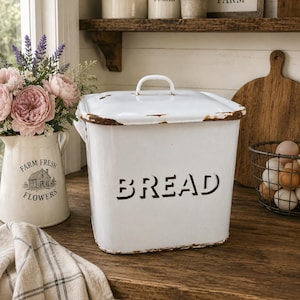 May include: A white bread bin with the word "BREAD" in black lettering. The bin has a handle on the lid and is on a wooden countertop. A vase of flowers and a checkered tea towel are nearby.