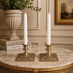 May include: A pair of matching brass candle holders with square, intricately engraved bases and cylindrical sockets, each holding a tall, slender, white taper candle. The metal has a polished gold-toned finish with detailed floral patterns.