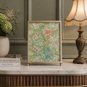 May include: A gold-coloured picture frame displaying a floral print with green leaves and red and blue flowers. The frame sits on a marble-topped table, beside a lamp and a vase of flowers.