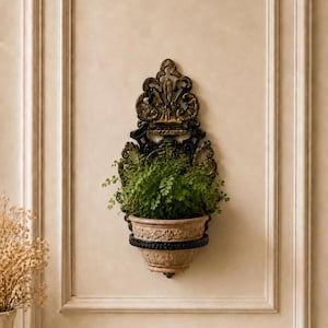 May include: A wall-mounted planter featuring an ornate black and gold metal frame with intricate scrollwork and a decorative crest. The planter holds a beige ceramic pot with a floral relief pattern, containing a lush green leafy plant.