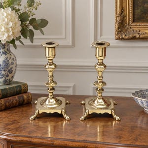 Peut inclure: Une paire de bougeoirs en laiton poli dot&eacute;s d'une tige haute et fine avec plusieurs anneaux d&eacute;coratifs et une base carr&eacute;e soutenue par quatre petits pieds incurv&eacute;s. La surface m&eacute;tallique est r&eacute;fl&eacute;chissante et lisse, avec une finition dor&eacute;e.