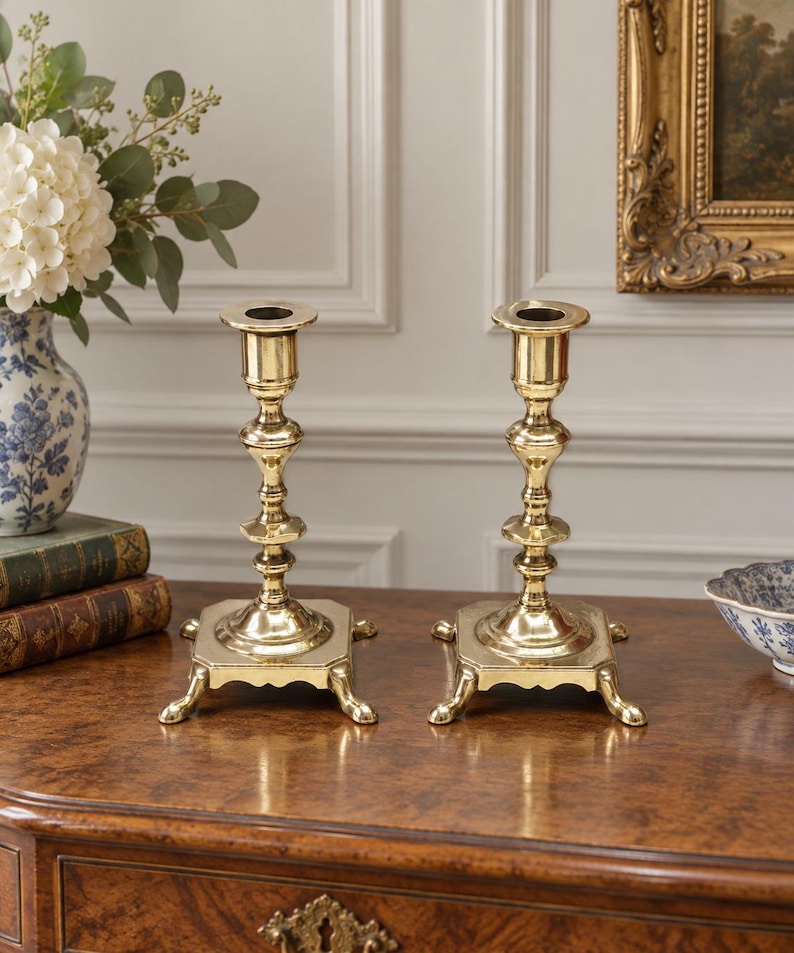 Peut inclure: Une paire de bougeoirs en laiton poli dot&eacute;s d'une tige haute et fine avec plusieurs anneaux d&eacute;coratifs et une base carr&eacute;e soutenue par quatre petits pieds incurv&eacute;s. La surface m&eacute;tallique est r&eacute;fl&eacute;chissante et lisse, avec une finition dor&eacute;e.