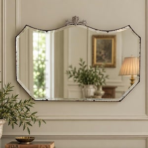 May include: A wall mirror with a scalloped, multi-faceted frame and a dark, weathered edge finish. The top centre features a decorative silver-toned metal crest. The glass surface is clear and reflects a room interior with a lamp and a plant.