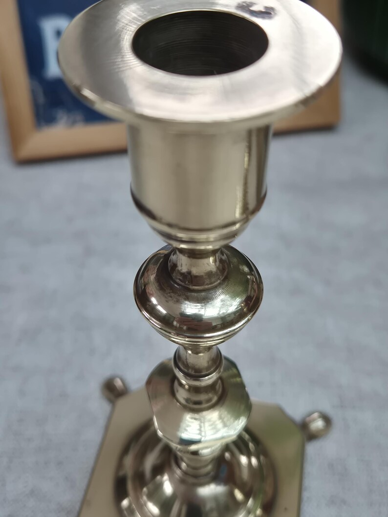 Peut inclure: Un bougeoir en m&eacute;tal argent&eacute; poli dot&eacute; d'une ouverture circulaire sur le dessus, d'une tige fine avec des n&oelig;uds d&eacute;coratifs arrondis et facett&eacute;s, et d'une base carr&eacute;e reposant sur de petits pieds arrondis &agrave; chaque coin de la structure.
