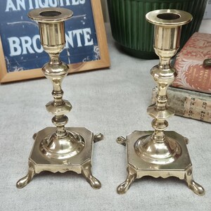 Peut inclure: Une paire de bougeoirs en laiton poli dot&eacute;s d'une tige haute et fine avec plusieurs anneaux d&eacute;coratifs et un sommet &eacute;vas&eacute;. Chaque bougeoir repose sur une base carr&eacute;e soutenue par quatre petits pieds incurv&eacute;s, offrant une base stable.